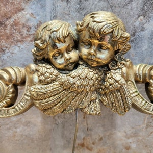 May include: Two gold cherub angels with wings, one with its arm around the other, are depicted in a close-up image. The angels are made of a metallic material and have a detailed, ornate design.