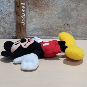 K&ouml;nnte beinhalten: Ein Stofftier von Mickey Mouse, das auf der Seite liegt. Das Spielzeug hat einen schwarzen Kopf und Ohren, wei&szlig;e Handschuhe, einen roten Torso mit wei&szlig;en Kn&ouml;pfen und gelbe Beine. Ein Holzlineal mit dem Text "Jodamel" befindet sich im Hintergrund.