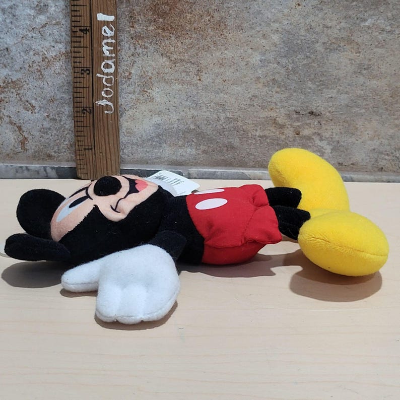 K&ouml;nnte beinhalten: Ein Stofftier von Mickey Mouse, das auf der Seite liegt. Das Spielzeug hat einen schwarzen Kopf und Ohren, wei&szlig;e Handschuhe, einen roten Torso mit wei&szlig;en Kn&ouml;pfen und gelbe Beine. Ein Holzlineal mit dem Text "Jodamel" befindet sich im Hintergrund.