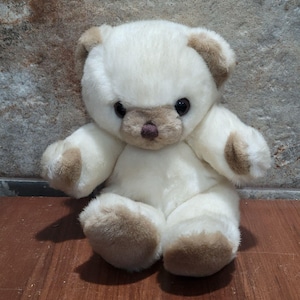 Può includere: Un orsacchiotto di peluche con pelliccia color crema e accenti marrone chiaro su orecchie, muso, zampe e piedi. Il peluche presenta grandi occhi scuri e rotondi e un piccolo naso marrone scuro, seduto in posizione eretta con pelo morbido.