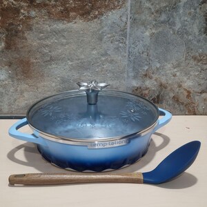 Pode incluir: Uma caçarola Temptations azul clara com tampa de vidro e uma pega prateada em forma de flor. O prato tem pegas azuis claras e uma base degradê azul escura. Uma colher de silicone azul com cabo de madeira está ao lado.