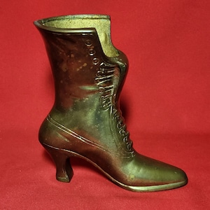May include: A gold-toned metal boot-shaped vase with a high heel. The boot has a lace-up design and is detailed with stitching.