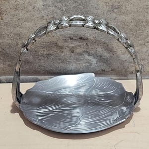 May include: A silver-colored metal basket with a handle adorned with floral details. The basket's base is shaped like a leaf with detailed veining. The handle is arched, and the overall design is ornate and decorative.