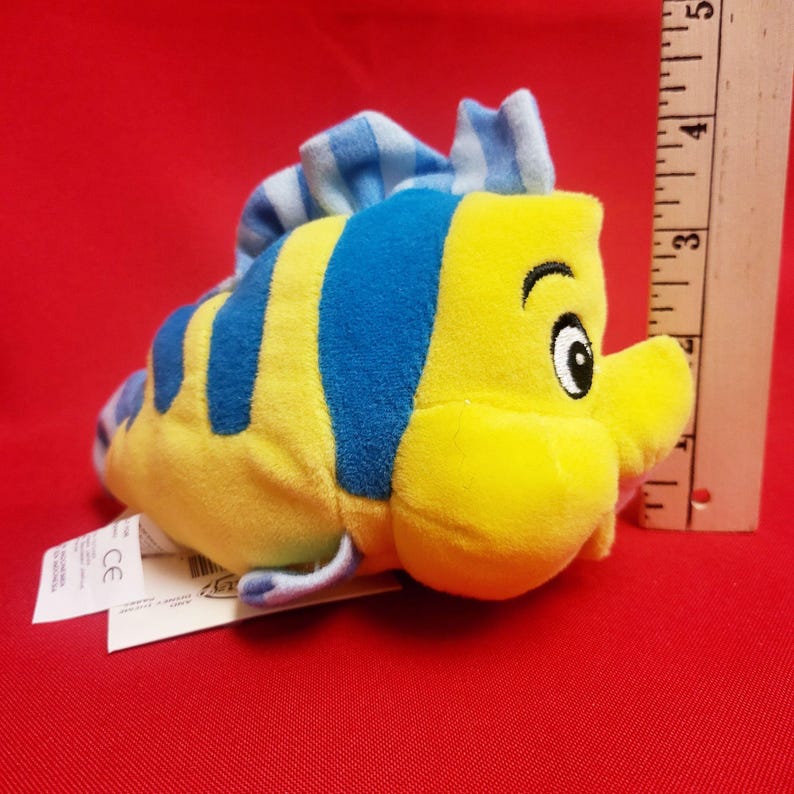 K&ouml;nnte beinhalten: Ein Pl&uuml;schtier der Figur Fabius aus Arielle, die Meerjungfrau. Das Spielzeug ist gelb mit blauen Streifen und einer blauen Flosse. Das Spielzeug ist etwa 10 cm lang.