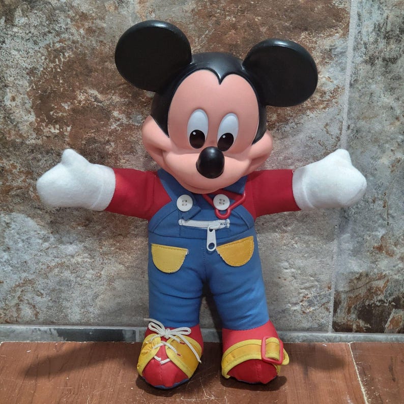 May include: A plush Mickey Mouse doll with black ears, a red shirt, blue overalls, and red shoes with yellow accents. The doll has white gloves and is posed with arms outstretched.