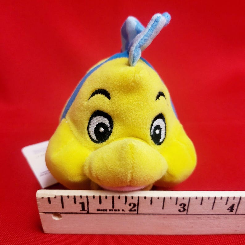 K&ouml;nnte beinhalten: Ein gelbes Pl&uuml;schfisch-Spielzeug mit blauen Akzenten, mit gro&szlig;en schwarzen Augen und einem l&auml;chelnden Gesichtsausdruck. Das Spielzeug ist etwa 7,6 cm lang und vor rotem Hintergrund.