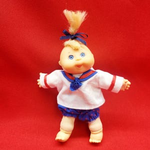 May include: A small, blonde haired doll wearing a white sailor shirt with red trim, a blue sailor collar, and blue and red polka dot shorts. The doll has blue eyes and a smiling face.