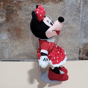 K&ouml;nnte beinhalten: Eine Minnie Mouse Pl&uuml;schpuppe mit rotem Kleid mit wei&szlig;en Herzakzenten, passender Schleife und roten Schuhen. Die Puppe hat einen schwarzen Kopf und Ohren, ein rosa Gesicht und wei&szlig;e Handschuhe. Die Puppe steht auf einem Metallst&auml;nder.