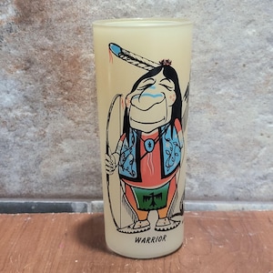 Op de afbeelding: Een geel glas met een cartoonillustratie van een indiaanse krijger die een boog en pijl vasthoudt. De krijger draagt een rood shirt, een blauw vest en een groene kilt. Het glas heeft het woord "WARRIOR" onderaan gedrukt.
