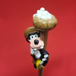 Puede incluir: Una figurita de personaje de dibujos animados de un perro con un sombrero marrón, trepando por un poste de madera con un nido de pájaro en la parte superior. El nido está lleno de huevos blancos.
