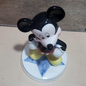 Puede incluir: Una figurita de cer&aacute;mica de Mickey Mouse de pie sobre una estrella azul. Mickey lleva sus pantalones cortos rojos caracter&iacute;sticos con lunares blancos, zapatos amarillos y guantes blancos. Tiene una nariz negra y orejas negras grandes.