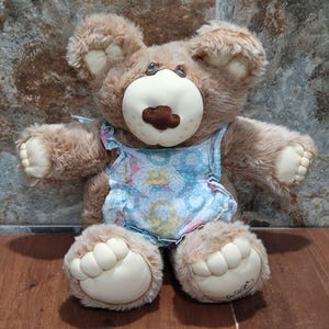 Op de afbeelding: Een bruine pluche teddybeer die een blauwe en witte bloemenjurk draagt. De beer heeft een bruine neus en grote, bruine ogen.
