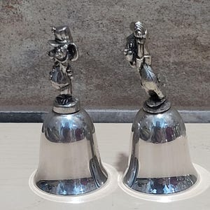 Pu&ograve; includere: Due campane color argento con figurine di personaggi dei cartoni animati in cima. Le campane hanno una base svasata e una superficie liscia e riflettente. Le figurine dettagliate aggiungono un tocco stravagante alle campane decorative.