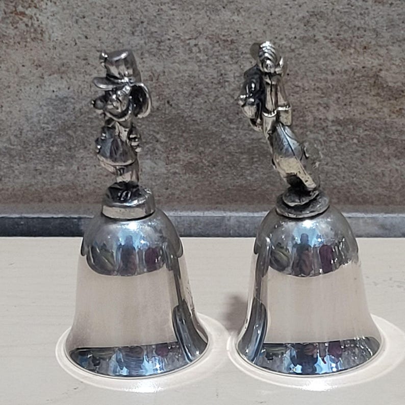 Pu&ograve; includere: Due campane color argento con figurine di personaggi dei cartoni animati in cima. Le campane hanno una base svasata e una superficie liscia e riflettente. Le figurine dettagliate aggiungono un tocco stravagante alle campane decorative.