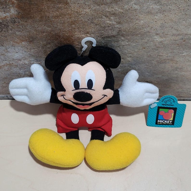 K&ouml;nnte beinhalten: Ein Mickey Mouse Pl&uuml;schtier mit schwarzen Ohren, einem schwarzen K&ouml;rper, roten Shorts und gelben Schuhen. Das Spielzeug hat wei&szlig;e Handschuhe und ein l&auml;chelndes Gesicht. Ein blaues Etikett mit "Mickey Unlimited" ist angebracht.