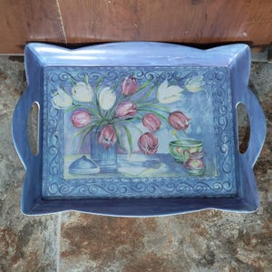 May include: A rectangular, blue serving tray with handles. The tray features a floral painting of pink and white tulips in a vase, with additional decorative elements. The tray is made of a durable material and is suitable for serving food and drinks.