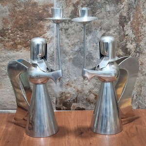Può includere: Due candelabri argentati a forma di angelo. Ogni figura di angelo tiene una coppa alta per candela. Gli angeli hanno ali stilizzate e gonne coniche. Lo sfondo è marrone e grigio screziato.
