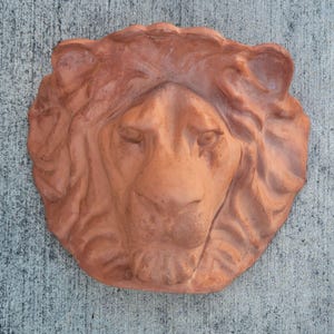Puede incluir: Escultura de cabeza de león de terracota con rasgos faciales detallados y una melena texturizada. La escultura es de color marrón rojizo cálido y probablemente está destinada a la exhibición al aire libre como adorno de jardín.