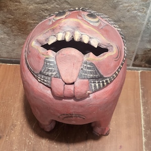 Pode incluir: Uma escultura de madeira esculpida à mão de uma criatura mítica com corpo arredondado, pintada num tom de vermelho suave. A figura apresenta uma boca larga e aberta que revela dentes esculpidos, uma língua proeminente e detalhes pretos.