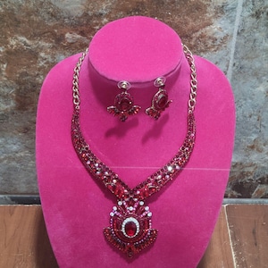 May include: A red jewelry set featuring a necklace and matching earrings. The necklace has a large pendant with a central oval-shaped red stone, surrounded by smaller red and white stones. The earrings are small, with a similar design. The set is displayed on a pink mannequin.