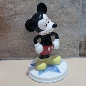 Puede incluir: Una figurita de cer&aacute;mica de Mickey Mouse de pie sobre una base azul en forma de estrella. Mickey lleva sus pantalones cortos rojos caracter&iacute;sticos con dos botones negros y zapatos amarillos. Tiene una cara blanca y negra con orejas grandes y mira hacia la derecha.