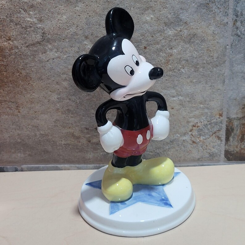 Puede incluir: Una figurita de cer&aacute;mica de Mickey Mouse de pie sobre una base azul en forma de estrella. Mickey lleva sus pantalones cortos rojos caracter&iacute;sticos con dos botones negros y zapatos amarillos. Tiene una cara blanca y negra con orejas grandes y mira hacia la derecha.