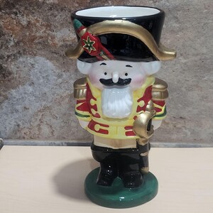 May include: A ceramic nutcracker figurine in a soldier's uniform. The figurine has a black hat with gold trim, a yellow jacket with red and gold accents, and black trousers. It stands on a green base and holds a gold sword.