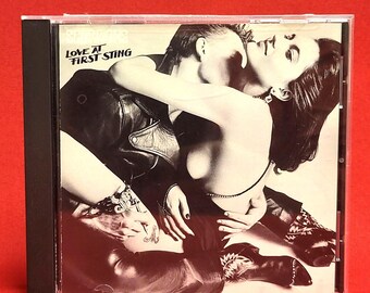 Puede incluir: Caja de CD con la portada del álbum "Love at First Sting". La imagen es una fotografía en blanco y negro de una pareja abrazándose. El título del álbum está impreso en letras negras en la parte superior izquierda.