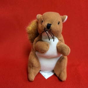 May include: A plush squirrel toy with brown fur, a white belly, and a fluffy tail. The squirrel has a black nose and whiskers. The toy is sitting on a red surface.