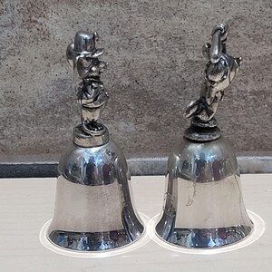 Pu&ograve; includere: Due campane color argento. Una campana ha la figura di una persona con cappello e abito. L'altra campana ha una figura simile a un uccello. Entrambe le campane hanno una base svasata e un manico in alto.