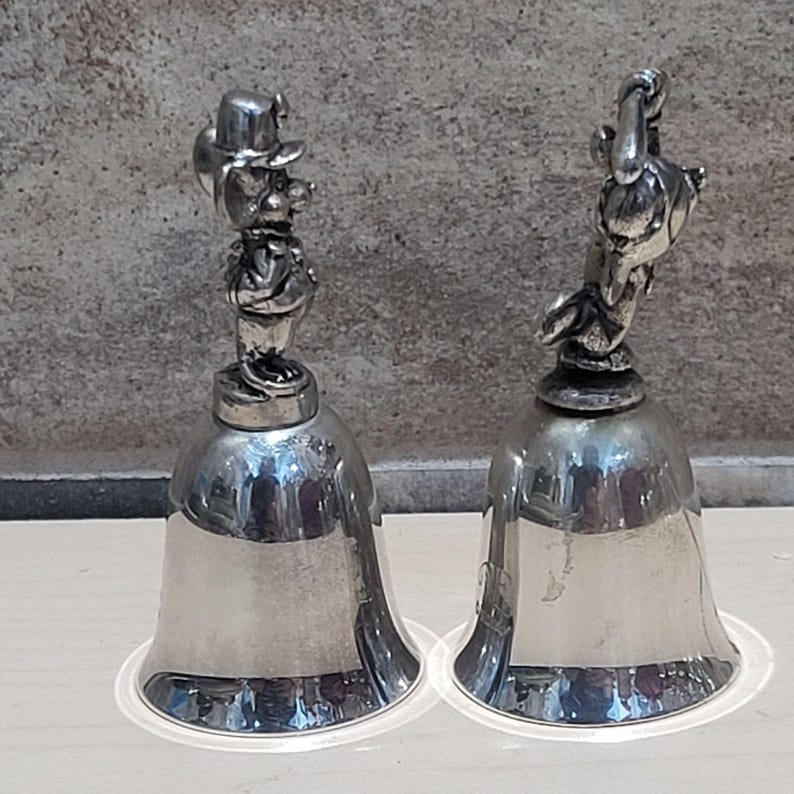 Pu&ograve; includere: Due campane color argento. Una campana ha la figura di una persona con cappello e abito. L'altra campana ha una figura simile a un uccello. Entrambe le campane hanno una base svasata e un manico in alto.