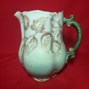 May include: A light green ceramic pitcher with a gold leaf design and a green handle. The pitcher has a scalloped rim and a rounded body.