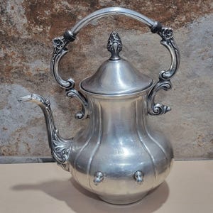 May include: A silver teapot with an ornate handle and lid. The teapot has a rounded body with a decorative design.