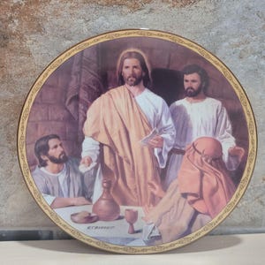 May include: A decorative round plate featuring a religious scene with several figures. The central figure is depicted in a white robe and tan shawl, surrounded by other figures. The plate has a gold border with an ornate design.