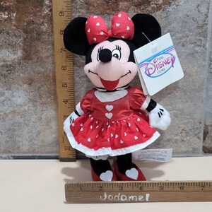K&ouml;nnte beinhalten: Minnie Maus Pl&uuml;schpuppe, mit rotem Kleid mit wei&szlig;en Herzakzenten, passender Schleife und schwarzen Ohren. Die Puppe hat wei&szlig;e Handschuhe und Schuhe mit Herzdetails. Ein Disney Store-Etikett ist angebracht.