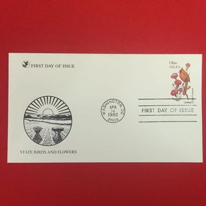 Pode incluir: Um envelope de primeiro dia de emissão branco com uma ilustração circular a preto e branco de um campo com feixes de trigo e um sol nascente. Inclui um selo de 20 centavos de Ohio com um cardeal e cravos vermelhos, e um carimbo.