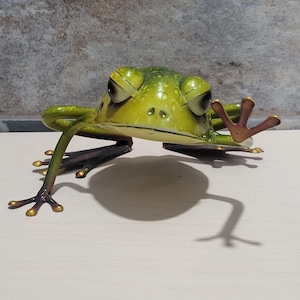 May include: A metal frog sculpture in shades of green and gold. The frog has large, expressive eyes and outstretched limbs, with a detailed texture. The sculpture is positioned on a light-colored surface, casting a shadow.