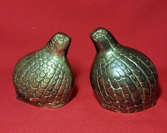 May include: Two gold-colored metal figurines of quail birds. The birds are facing each other and have detailed feather patterns.