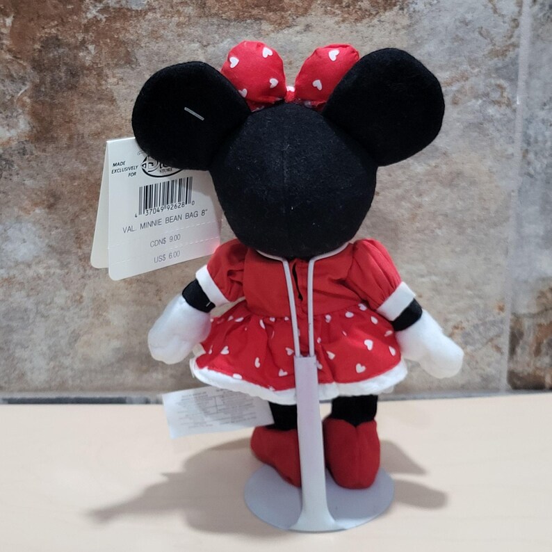 K&ouml;nnte beinhalten: Minnie Mouse Bean Bag Pl&uuml;schtier, mit einem roten Kleid mit wei&szlig;en Herzakzenten, einer passenden Schleife und schwarzen Ohren. Das Spielzeug hat wei&szlig;e Handschuhe und rote Schuhe. Ein Etikett ist am Spielzeug befestigt.