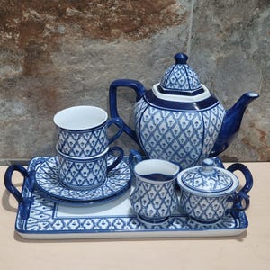 May include: A blue and white porcelain tea set with a teapot, two teacups, saucers, a creamer, a sugar bowl, and a serving tray. The set features a repeating geometric pattern. The teapot has a curved spout and handle.