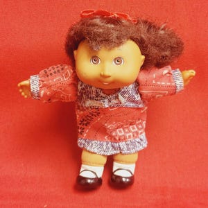 Puede incluir: Una pequeña muñeca Cabbage Patch Kid con cabello castaño, vestida con un vestido estampado en rojo y azul y zapatos negros. La muñeca tiene una tez rosada y ojos marrones.
