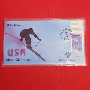 May include: A blue and white commemorative envelope with a stamp depicting a skier in silhouette. The envelope is for the 1994 Winter Olympics in Salt Lake City, Utah. The text on the envelope reads "Alpine Skiing" and "USA Winter Olympics".