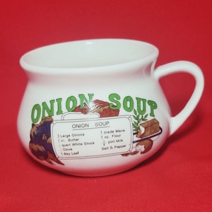 Puede incluir: Taza de sopa de cerámica blanca con una ilustración verde y marrón de un tazón de sopa de cebolla y las palabras "ONION SOUP" impresas en verde. La taza tiene una receta de sopa de cebolla impresa en el lateral, incluyendo ingredientes como cebollas, mantequilla, caldo blanco, macis, harina, leche, sal y pimienta.