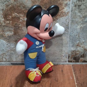 May include: A Mickey Mouse plush toy, featuring a black head and ears, a pink face, and white gloves. Mickey wears a red shirt, blue overalls with yellow accents, and red shoes with yellow trim. The toy is standing.