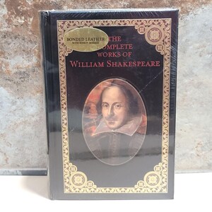 May include: A black book titled "The Complete Works of William Shakespeare" with a portrait of Shakespeare. The book is wrapped in plastic and has gold-colored decorative borders and text. The cover reads "Bonded Leather with Ribbon Marker."