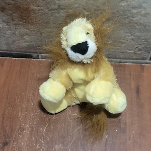 Puede incluir: Un león de peluche con pelaje suave de color amarillo claro, una melena desgreñada de color marrón oscuro y una nariz negra. El animal de peluche está sentado con las patas delanteras hacia adelante y una cola larga en la base.