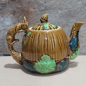 May include: A brown ceramic teapot with a squirrel handle and green leaf accents. The teapot is shaped like a barrel and has a lid with an acorn finial.