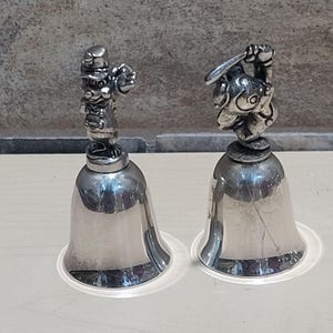 Pu&ograve; includere: Due campane color argento con figurine di personaggi dei cartoni animati sulla parte superiore. Una campana presenta una figura in uniforme da baseball, che tiene una mazza. L'altra campana ha una figura in uniforme da baseball, che tiene una palla da baseball. Le campane sono su basi di plastica trasparente.