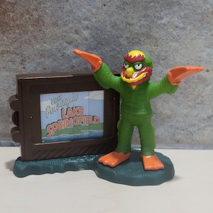 May include: A green monster toy with orange flippers and a red and yellow head. The monster is holding a brown sign that says "Visit Beautiful Lake Springfield".