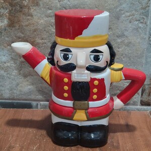 May include: A novelty ceramic teapot in the form of a nutcracker soldier. The teapot is predominantly red, with white, black, and yellow details. It has a red hat, a stern face with a black moustache, and a red uniform with gold epaulettes.